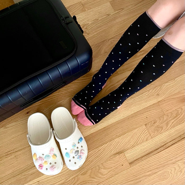 Knee-high black socks with colorful heart patterns, paired with white clogs, next to a suitcase on wooden floor.