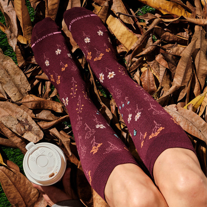 Knee-high burgundy compression socks with floral patterns, resting on dry leaves beside a coffee cup.