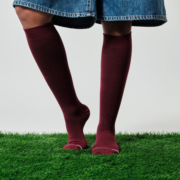 Knee-high compression socks in burgundy worn with a denim skirt, standing on green grass.