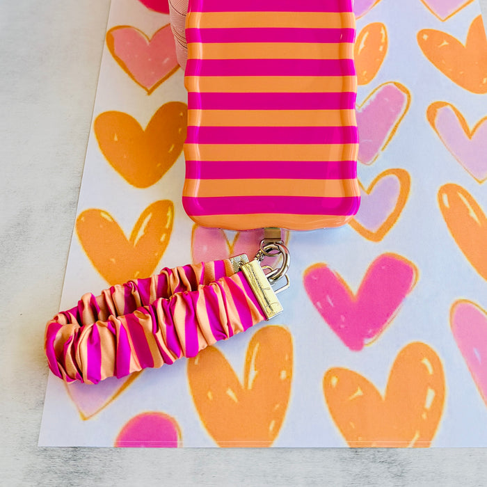 Pink and orange striped Candy Citrus Stripes scrunchie phone wrist strap clipped to the matching hot pink and citrus orange striped Wavy Luxe iPhone case, styled on a playful pink and orange heart print background.