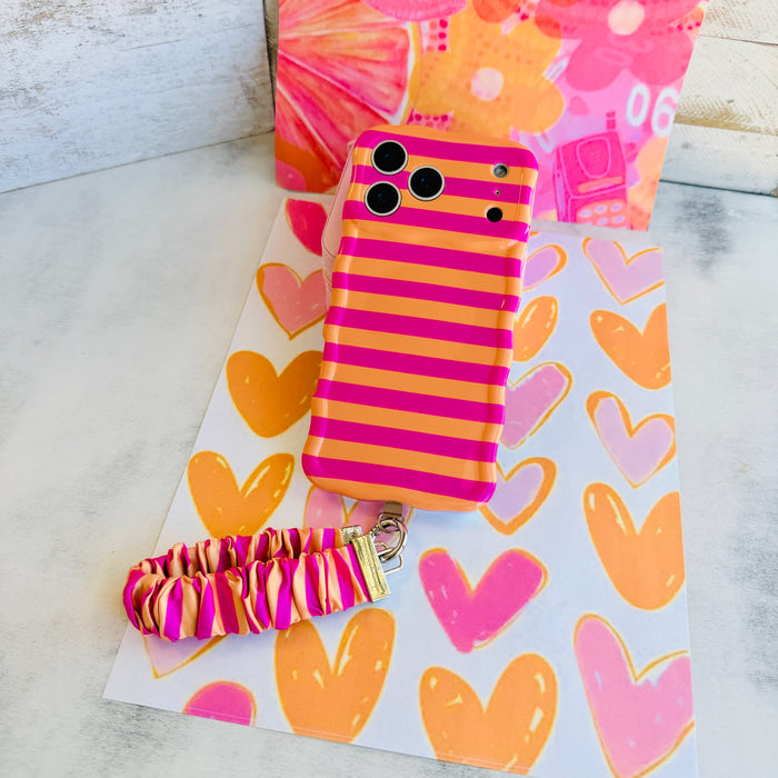 Pink and orange striped Candy Citrus Stripes scrunchie phone wrist strap clipped to the matching hot pink and citrus orange striped Wavy Luxe iPhone case, styled on a playful pink and orange heart print background.