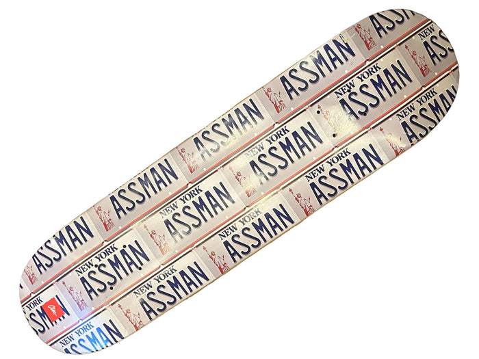 A skateboard deck featuring a repeating 'ASSMAN' and 'NEW YORK' license plate style pattern in blue, red, and white, photographed diagonally on a textured gold surface.