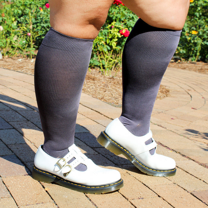 Wide calf knee-high compression socks in dark gray worn with white shoes, set against a garden background.