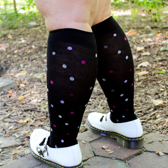 Knee-high compression socks with a black background and colorful polka dots, worn with white shoes on a natural surface.