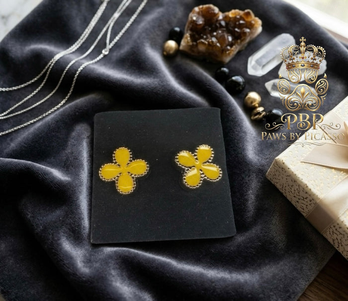 Yellow clover-shaped earrings on a black card with a dark fabric background and decorative elements.