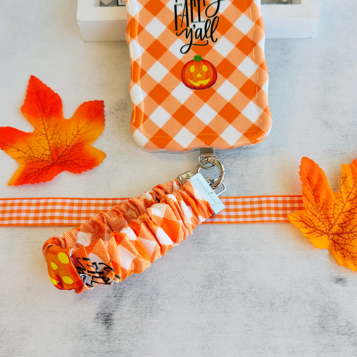 An orange gingham Happy Fall Y'all scrunchie phone wrist strap with a jack-o'-lantern and 'happy fall y'all' print, clipped to the matching Happy Fall Y'all Wavy Luxe iPhone case, styled with autumn maple leaves.