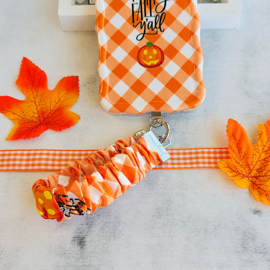 An orange gingham Happy Fall Y'all scrunchie phone wrist strap with a jack-o'-lantern and 'happy fall y'all' print, clipped to the matching Happy Fall Y'all Wavy Luxe iPhone case, styled with autumn maple leaves.