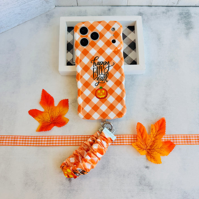 An orange gingham Happy Fall Y'all scrunchie phone wrist strap with a jack-o'-lantern and 'happy fall y'all' print, clipped to the matching Happy Fall Y'all Wavy Luxe iPhone case, styled with autumn maple leaves.