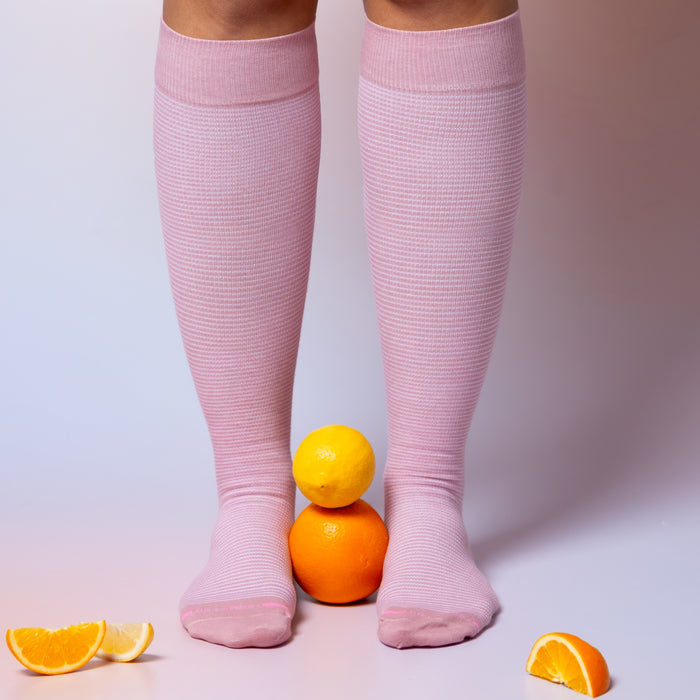 Knee-high pink compression socks with a microstripe pattern, positioned near stacked citrus fruits.