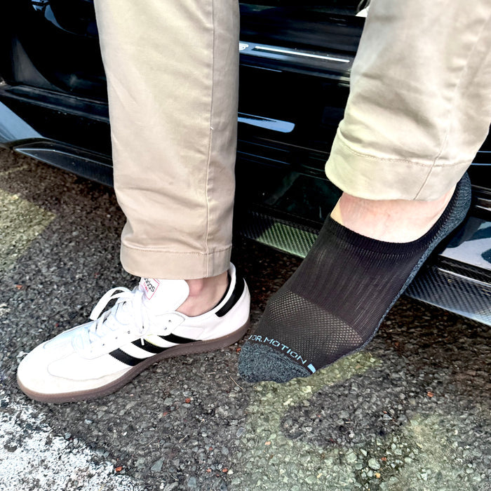 Black liner compression sock worn on one foot next to a white sneaker on a gravel surface.