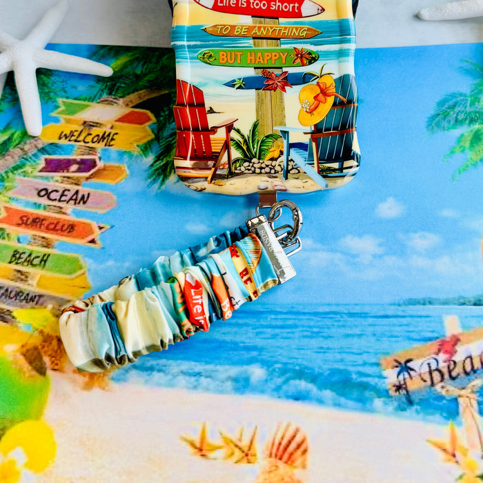 A colorful Seaside State Of Mind scrunchie phone wrist strap in a vintage beach print with Adirondack chairs, surfboard signposts, and palm trees, clipped to the matching Seaside State Of Mind Wavy Luxe iPhone case, styled on a tropical beach background with starfish and shells.