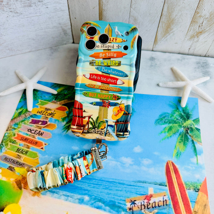 A colorful Seaside State Of Mind scrunchie phone wrist strap in a vintage beach print with Adirondack chairs, surfboard signposts, and palm trees, clipped to the matching Seaside State Of Mind Wavy Luxe iPhone case, styled on a tropical beach background with starfish and shells.
