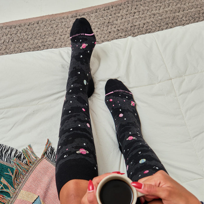 Knee-high black compression socks with a galaxy print, featuring planets and stars, worn on a bed.
