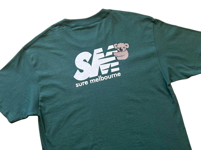 A dark green t-shirt laid flat on a brown wooden surface, featuring a white 'sure melbourne' logo and a small koala graphic on the back.