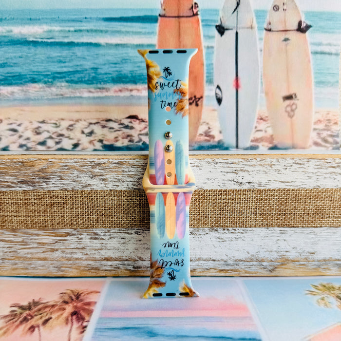 Sweet Summer silicone Apple Watch band with pastel pink, yellow, and blue surfboards, palm trees, and "sweet summer time" script on a soft watercolor beach scene, shown displayed and worn on Apple Watch against a coastal surfboard backdrop.