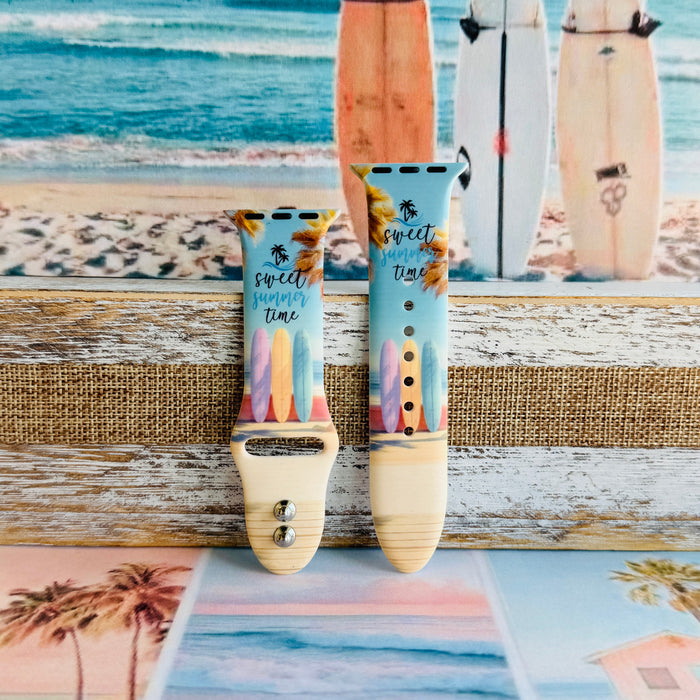 Sweet Summer silicone Apple Watch band with pastel pink, yellow, and blue surfboards, palm trees, and "sweet summer time" script on a soft watercolor beach scene, shown displayed and worn on Apple Watch against a coastal surfboard backdrop.