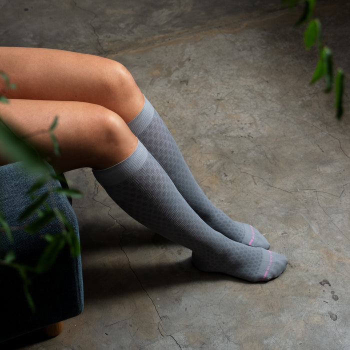 Knee-high compression socks in gray with a textured pattern and pink accents, worn by a seated person.